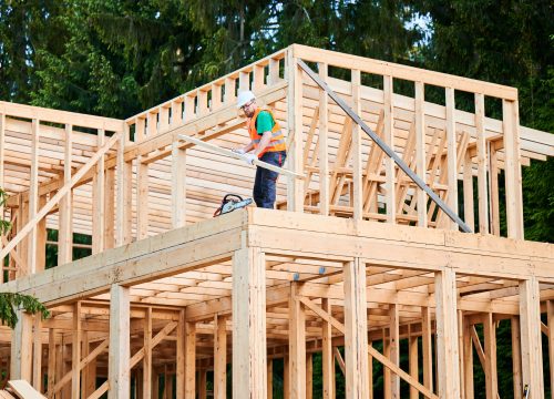 Carpenter constructing wooden-framed house near the woods. Man holding large joist, wearing work clothes and helmet. Concept of modern and eco-friendly building.